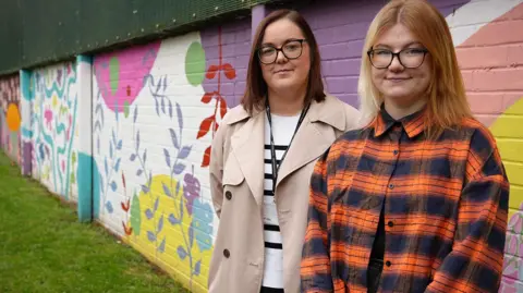 University of Wolverhampton Two women are stood in front of a wall which has colourful floral patterns painted on it.