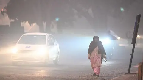 Getty Images A woman wearing a pink traditional Indian dress and a black scarf walks along the side of the road in the early hours of the morning in Karol Bagh, Delhi, as vehicles pass by with their lights on due to low visibility.