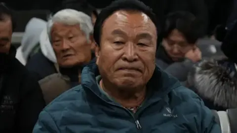 Maeng Gi-su wears a green puffa jacket and a sombre expression.