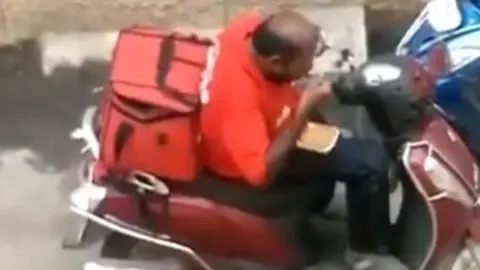 @Madan_Chikna Male Zomato delivery driver eating food from a takeaway container.