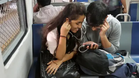 Getty Images This photo taken on March 7, 2017 shows Indian students watching a movie on their smartphone while commuting on a suburban train in Mumbai. Buyouts, mergers and quick exits -- as India's richest man shakes up the country's ultra-competitive mobile market, telecommunications companies are scrambling to either consolidate or cut their losses and run.