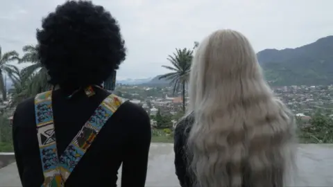 Olivier Assoua Two women, one with afro, another in a blonde wig, watch the city their backs turned to the camera
