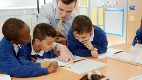 BBC Man teaches three secondary school boys in classroom at their desk with work