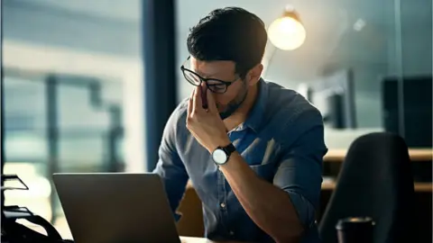 Getty Images A stressed man pinches the bridge of his nose in frustration in front of his laptop in this photo illustration