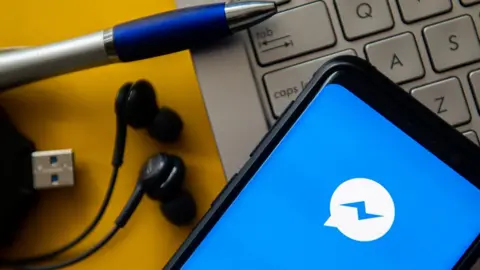 Getty Images The Facebook messenger logo is seen on a mobile phone lying on a laptop, next to some pens and earphones and a USB cable