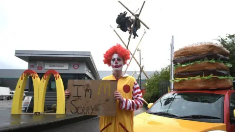 PA Media An Animal Rebellion protester, dressed as a clown, and protesters suspended from a bamboo structure outside a McDonalds distribution site in Hemel Hempstead