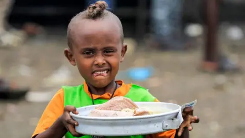 AFP A child walks with a tray carrying soup and traditional Injera Ethiopian sour flatbread as Ethiopian refugees of the Qemant ethnic group queue for food at a camp in the village of Basinga in Basunda district of Sudan's eastern Gedaref region on August 10, 2021.