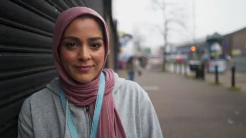 BBC Hana, a young woman wearing a hijab and lanyard, outside a shop on a road in Leeds