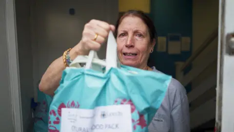BBC Janice, stood in the food hub doorway holding a blue bag full of food