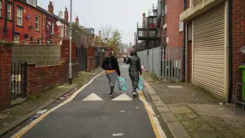 BBC Two women walk down a road holding blue bags full with food