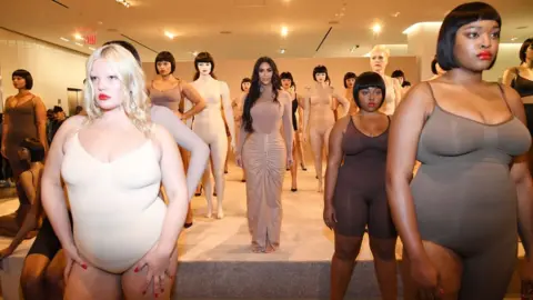 Getty Images Kim Kardashian, with dark hair down to her waist, stands among a diverse group of models at the launch of her shapewear brand, Skims, at a shop in New York in February 2020. The models in the foreground include a blonde, white woman with red lipstick wearing a white body suit and to her left there is a taller black woman with bobbed brown hair wearing a brown body suit and the same red lipstick. Behind Kim there are more models as well as mannequins, many of which have bobbed dark hair. Kim is wearing a long sleeved nude coloured top and floor length skirt that gathers in the middle.