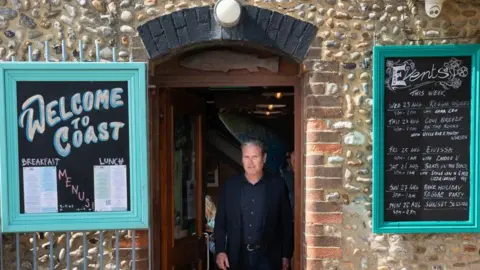 PA Media Labour leader Sir Keir Starmer visits Worthing in West Sussex. He is standing in the doorway of a cafe looking at the camera and wearing a dark blue shirt and suit jacket. Either side of him on the brick wall of the front of the cafe are black chalkboard signs advertising the breakfast and lunch menus to the left and then the events for the week to the right.