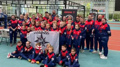 Compass Martial Arts More than 45 competitors, including children, in red and blue training uniforms, pose holding a white Compass Martial Arts flag at the competition in Frankfurt, Germany.