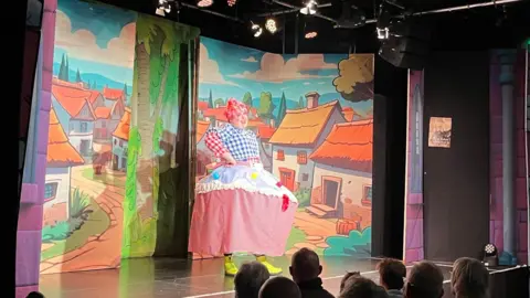 George Carden/BBC Alex wearing an outfit which looks like a cupcake up on stage, including a blue, white and red chequered blouse, pink wig and a huge pink skirt with a hoop. He is standing on stage in front of scenery that depicts a cartoon village. There are a few audience members looking on in the foreground.