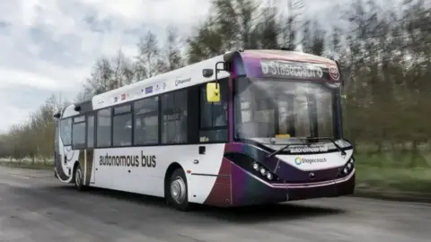 PA Media A purple and white Stagecoach bus marked "autonomous bus" on the front and side drives along a tarmac road with grass and trees on the verge. 