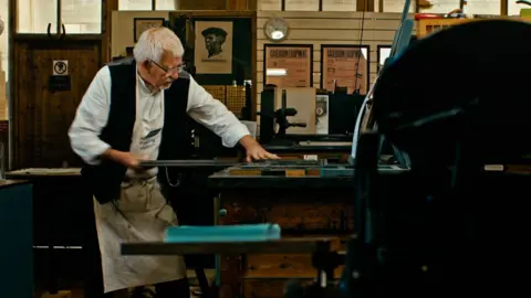 Norwich Printing Museum A man with white hair and glasses. He is wearing a white shift with a black sleeveless jacket and an apron around his waist. He is pulling a metal bar away from a table. He is focussed on the table to his left. In the background, the walls are busy with artwork. To the far left in the foreground, there is the silhouette of machinery. 