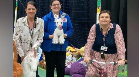 Short Wood Primary School Three women from the school are standing in a sports hall in front of a football goal. One is holding a white and pink stuffed unicorn toy. The next is holding a small white reindeer stuffed animal, and the final woman is straddling a pink and white children's bike