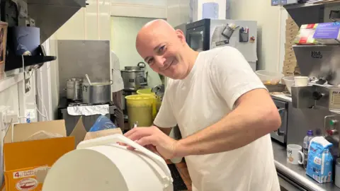 BBC Ronnie Christodoulou working at the back of his chip shop. He is wearing a white t-shirt and is bald. 