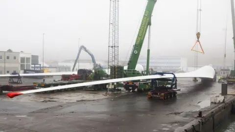 Vestas A large single wind turbine blade is lifted by crane onto specially designed trucks on a foggy dockside
