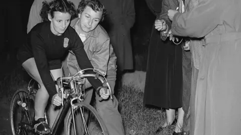 George W. Hales/Fox Photos/Getty Images A black and white photo of Eileen Sheridan setting off on a record-breaking run from London to Oxford. She is wearing a dark top and shorts, as crowds stand around her
