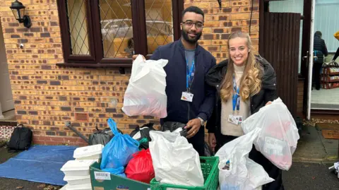 Volunteers pictured with bags of food. A man wearing a blue jacket and glasses holds up a bag of food items. A woman next to him has long blonde hair, a beige jumper and black coat. Both wear work identification and are smiling at the camera.