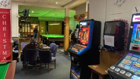 A view across a social club with the corner of a snooker table to the left and slot machines to the right. A red banner has white text reading "Merry Christmas". In the middle-distance a woman sits in a comfortable bar chair at a table. In the background a man can be seen standing next to a snooker table.