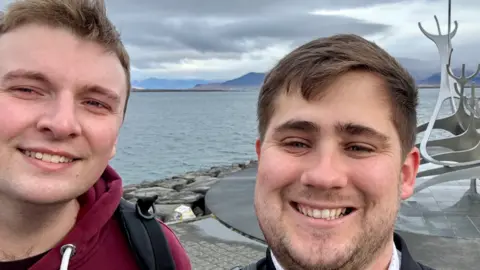 Zak Nelson Elliot Griffiths and Zak Nelson facing the camera and smiling in a selfie in front an abstract metal sculpture with water and mountains in the background