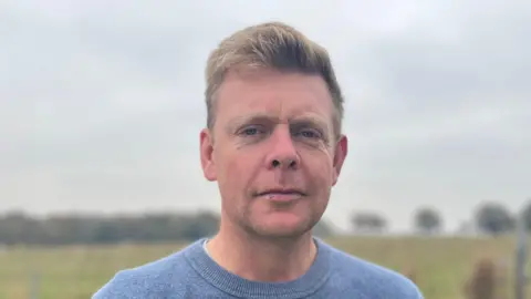 BBC Richard Baugh is pictured. He has blonde hair and white skin and is wearing a blue crew neck jumper.