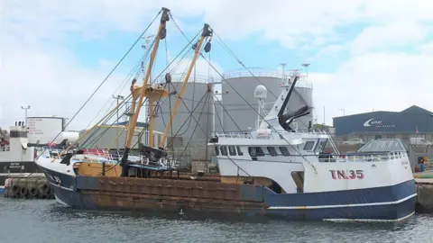 Marine Accident Investigation Branch Scallop-trawler Olivia Jean in a harbour, with blue hull and yellow rigging, with industrial buildings in the background