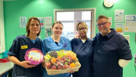 BBC Matt stands on the right next to three nurses from ward 408 of the Queen Elizabeth Hospital. One nurse is holding a box of chocolates and another holds a fruit hamper.