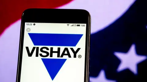 Getty Images The Vishay Intertechnology Semiconductor manufacturing company logo displayed on a smartphone. The logo is a blue triangle pointing downwards on top of a white background. The triangle is broken across the middle with the word VISHAY in black capital letters. A blurred American flag is in the background behind the phone.