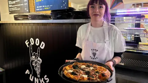 SOSE A young woman with long purple hair holds out a pizza on a plate in front of a food counter and kitchen