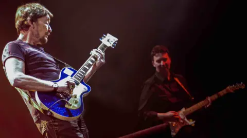 Paul Casey Guitarists Chris Rea, dressed in dark t-shirt and playing a blue guitar, and Paul Casey, in a dark shirt and playing a fender , play together on a darkly lit stage