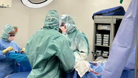 BBC Three people wearing green scrubs stand over a surgical table. There is a person on the table who cannot be seen in the image. They are performing hip surgery. 