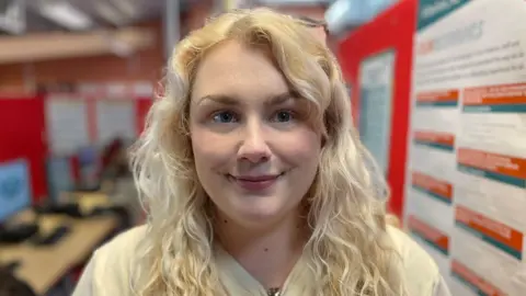 Laura Foster/BBC Charlotte wears her blonde, curly hair down with a white top. She is standing in a room with computer screens and red boards.