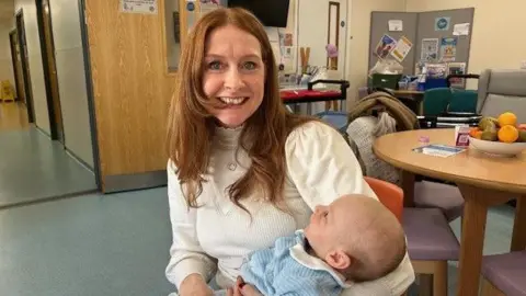 BBC A woman with red hair and a white top holding a baby in blue clothing.