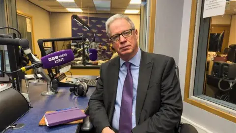 BBC Doug Bannister sits in a chair in the BBC Radio Kent studio with a branded microphone pointing towards him. He is looking at the camera, wearing glasses and a suit with a blue shirt and purple tie