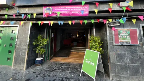 BBC Colourful bunting flags hang over an open doorway which has steps and a mobility ramp going into the cultural hun. There's a green billboard sign outside with two plants either side of the door. There's a pink banner up over the doorway which has the CCDP logo on it.