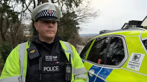 BBC Sgt Dan Ayrton from Surrey's Roads Policing Unit, in front of a police car.