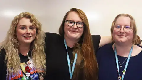Dominic Ward Three women stand against a beige wall with their arms around each others' shoulders. On the left, Charlotte has her blonde curly hair down and she is wearing a black band T-shirt. In the middle, Laurie has her red hair done and on one side and is wearing a black T-shirt and glasses. Stephanie is wearing a navy blue top and heart necklace, and pale glasses. They are all looking at the camera and smiling.