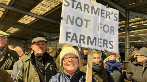 BBC Farmer Nicky Robinson from Ledbury holds a placard saying "Starmer's not for farmers". She stands in a crowd at an auction centre. 