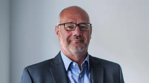 Guernsey Energy Darryl Goddard. He is wearing a grey suit, blue shirt and glasses. He has short grey hair and stubble. 