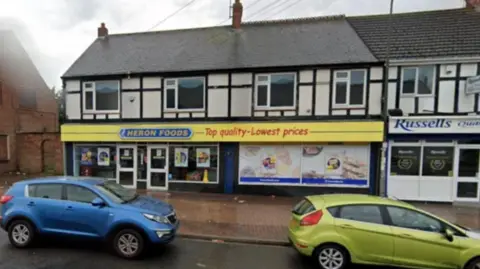 Google Heron Foods is in a mock Tudor row of terraced houses and has a yellow shop front with a blue sign. It has been raining and two cars are parked in front of the shop.