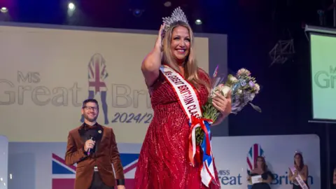 Miss Great Britain Debbie Hughes is on a stage, there is a sign on the wall behind her that says "Ms Great Britain 2024/2025". She is wearing a sparkly red long dress and a silver crown on her head. She has a sash on that says "Ms Great Britain Classic", and she is holding a bouquet of flowers, and has the other hand on the crown.