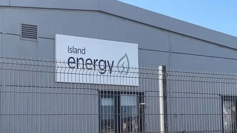 BBC A grey, plastic-clad industrial building with an Island Energy sign and logo on it. The logo has a mint green leaf icon with a power lightning flash in the middle. In front of the building is a high metal fence.