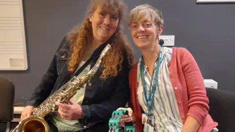 Absolute Music Trust Gaynor has long curly red hair with a fringe, styled in a topknot. She is sat, smiling at the camera, wearing a dark denim jacket and a pale green t shirt underneath and holds a saxophone. Lorna Farmer sits to her right, she is also smiling at the camera. Lorna has short blonde hair with a long fringe framing her face, she wears a blue striped blouse with a coral pink cardigan and has a blue NHS lanyard around her neck and holds a turquoise coloured tambourine.