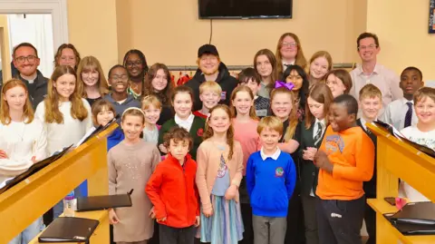 Lucy Taylor Photography Ed Sheeran standing in a room against a wall, surrounded by about 24 children. He is in the back row wearing a black cap and black jacket with red hair and bead and smiling. There are three other adults, also in the back row