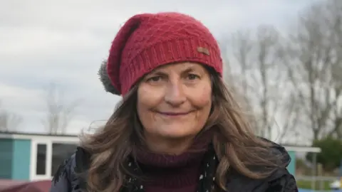 Jamie Niblock/BBC Woman stands on jetty and is wearing a red bobble hat, dark coat and red jumper. She has a black scarf with stars on it. Behind her are an array of boats.