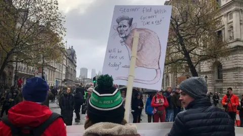 BBC Protestors carry banners including one with a cartoon image of Prime Minister Keir Starmer's head with a rat's body. The words on the sign read 'Starmer fled Welsh farmers "like a rat".  Will he face us today?'