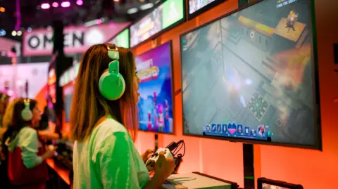 Getty Images A woman wearing a white T-shirt and over the ear headphones plays on the Xbox at a stand at a video games trade fair. She's holding the controller and looking at a large screen in front of her.